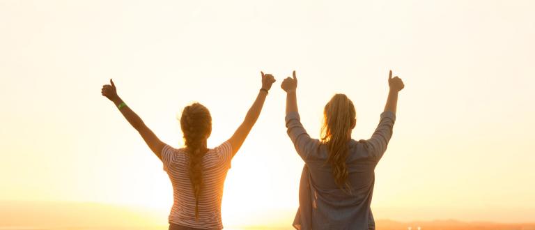 Two women with thumbs up in facing a sunset