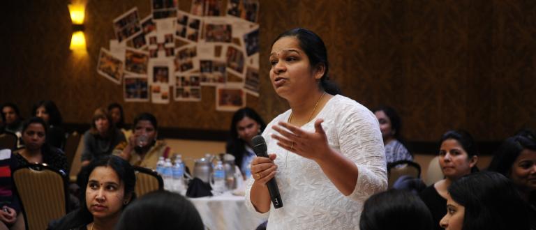Image of woman talking in a microphone, in a room full of women who work in high tech as consultants in the US. They are the best and brightest from universities in India. 