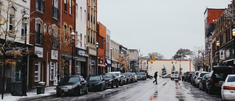 The image shows a snowy city street with parked cars and a person crossing the road, under an overcast sky. 