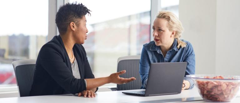 Deux femmes discutant à une table de conférence avec un ordinateur portable, engagées dans une conversation professionnelle.