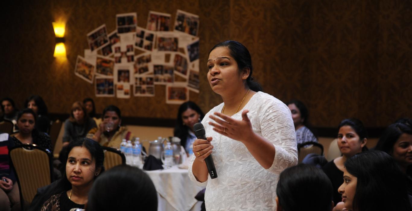 Image of woman talking in a microphone, in a room full of women who work in high tech as consultants in the US. They are the best and brightest from universities in India. 