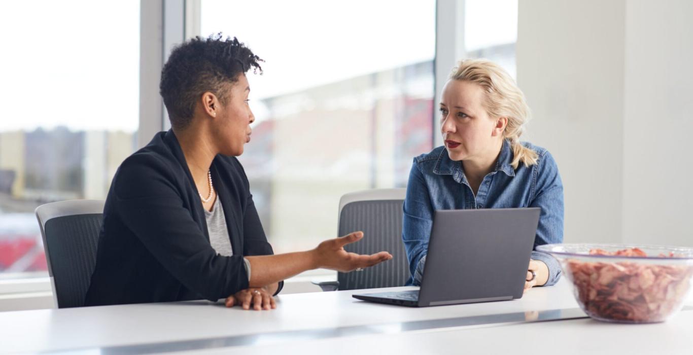 Deux femmes discutant à une table de conférence avec un ordinateur portable, engagées dans une conversation professionnelle.